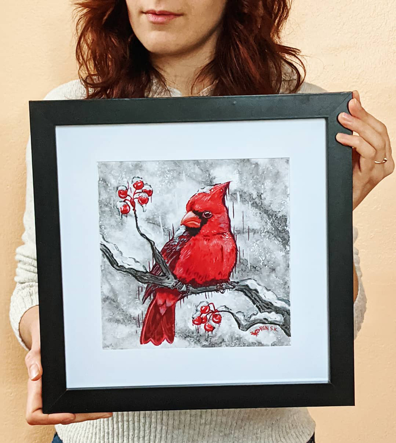 artist holding a framed portrait of a northern cardinal painted in gouache and sumi ink