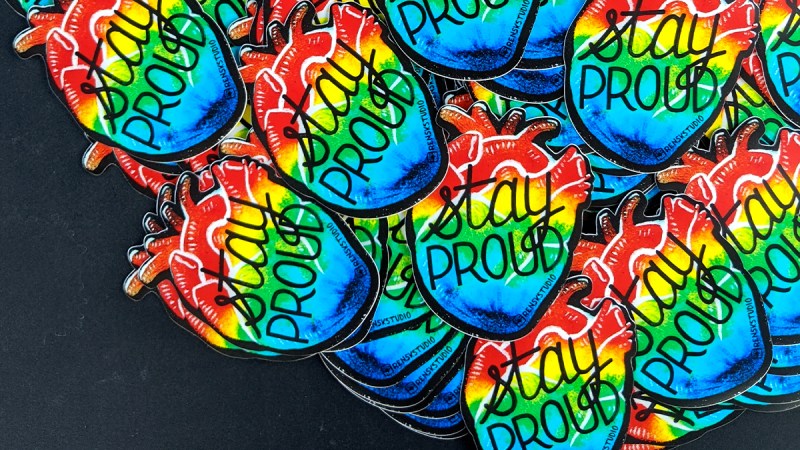 Pride rainbow stickers spread over a table, heart shaped that say "Stay Proud"