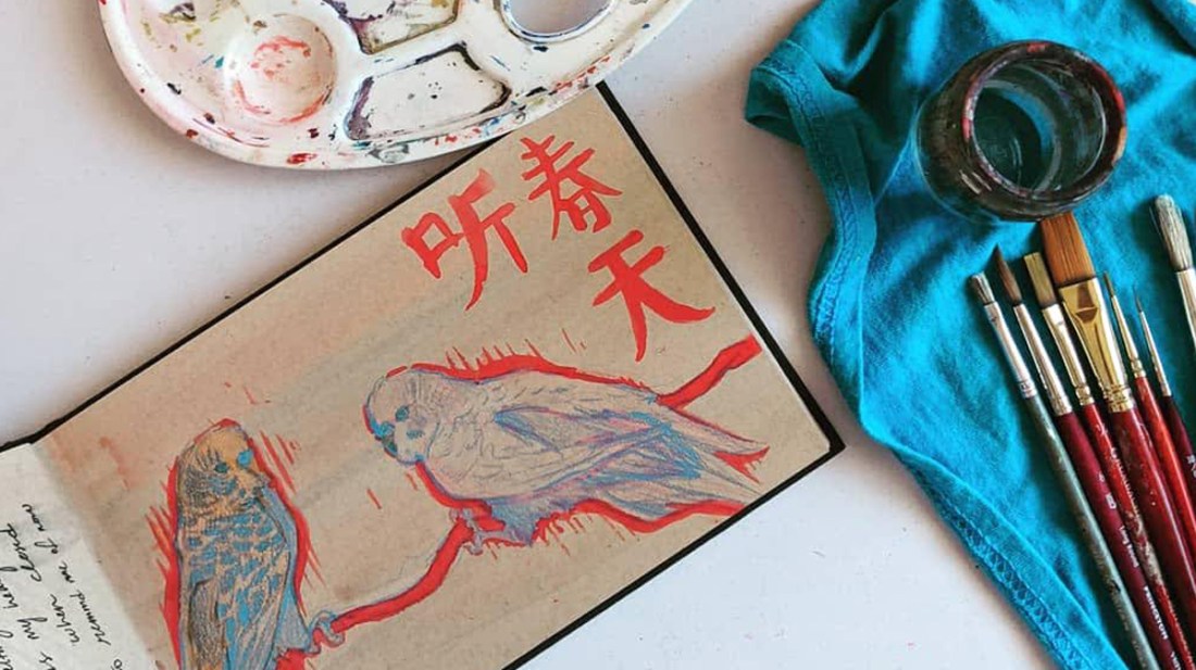 Desk with sketchbook open, colored pencils, brushes and palette. Sketchbook is open to a drawing of two budgies in red, blue, orange and pink.
