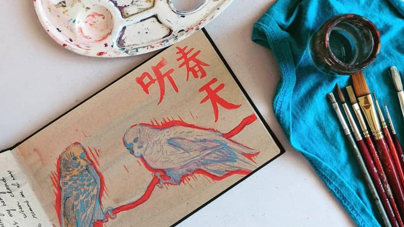Desk with sketchbook open, colored pencils, brushes and palette. Sketchbook is open to a drawing of two budgies in red, blue, orange and pink.