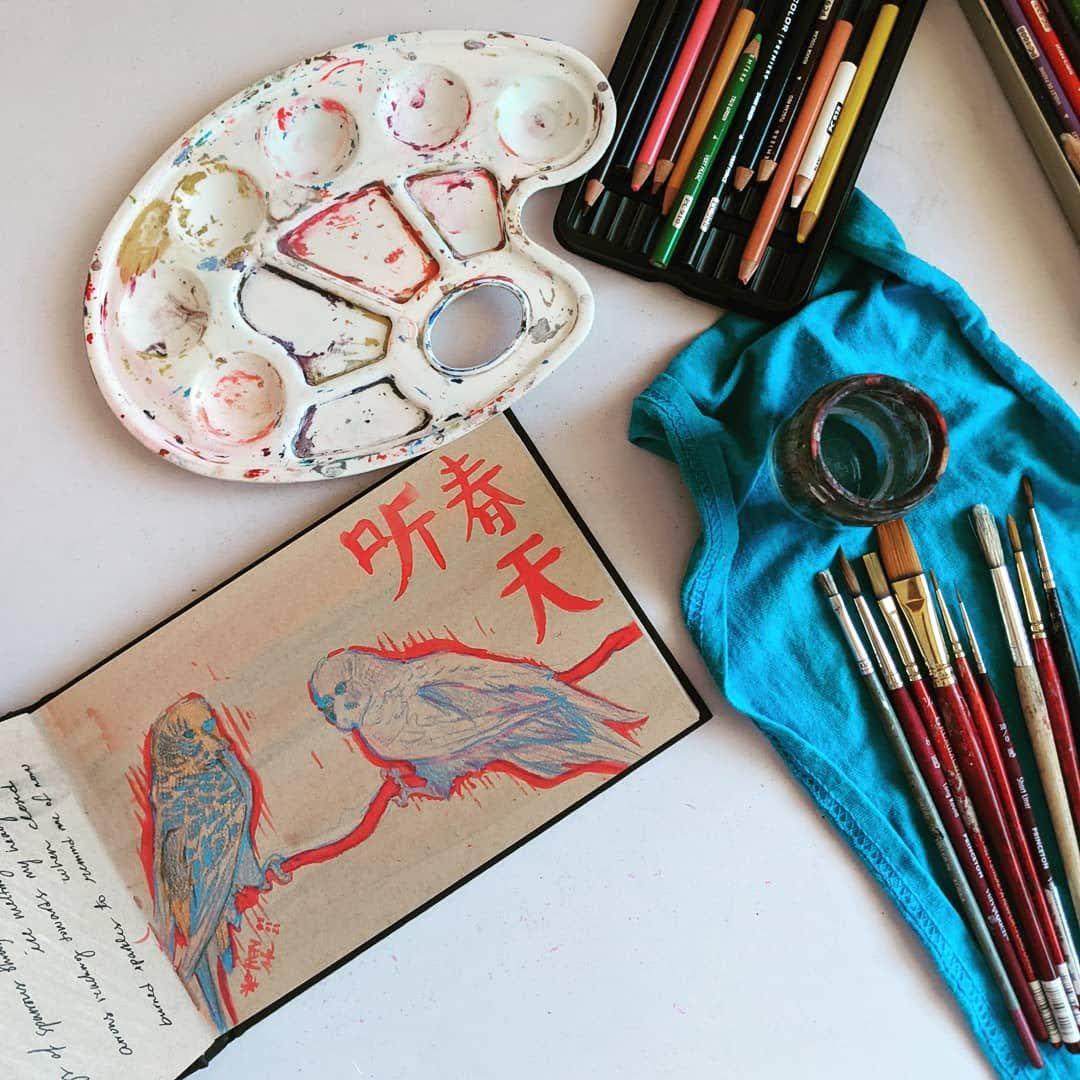 Desk with sketchbook open, colored pencils, brushes and palette. Sketchbook is open to a drawing of two budgies in red, blue, orange and pink.