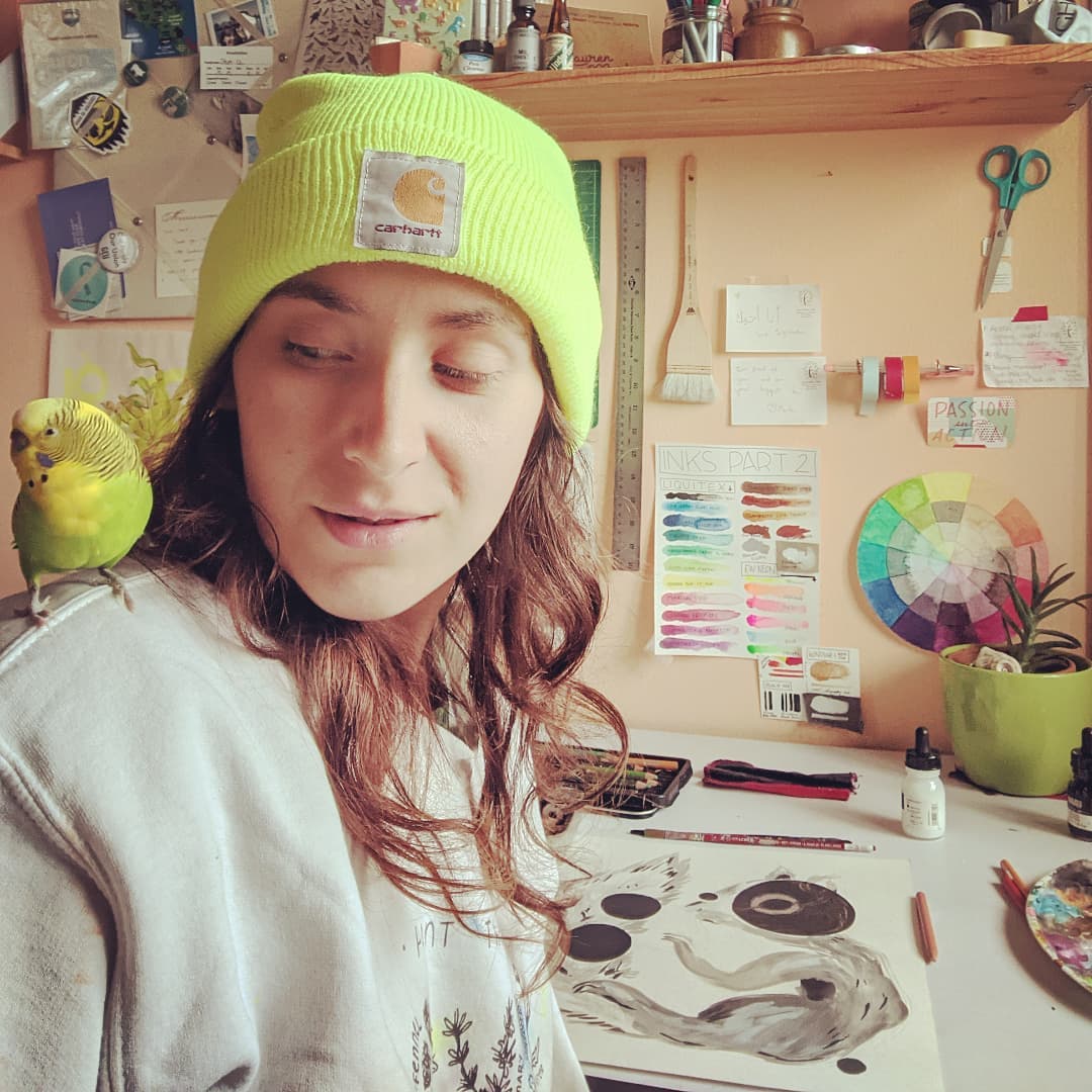 A photo of Ren at her drafting table with their lil parakeet supervisor, Opa Bird, on their shoulder