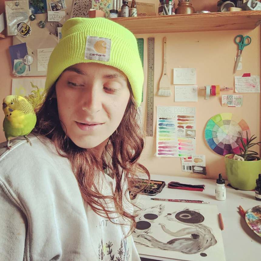 A photo of Ren at her drafting table with their lil parakeet supervisor, Opa Bird, on their shoulder