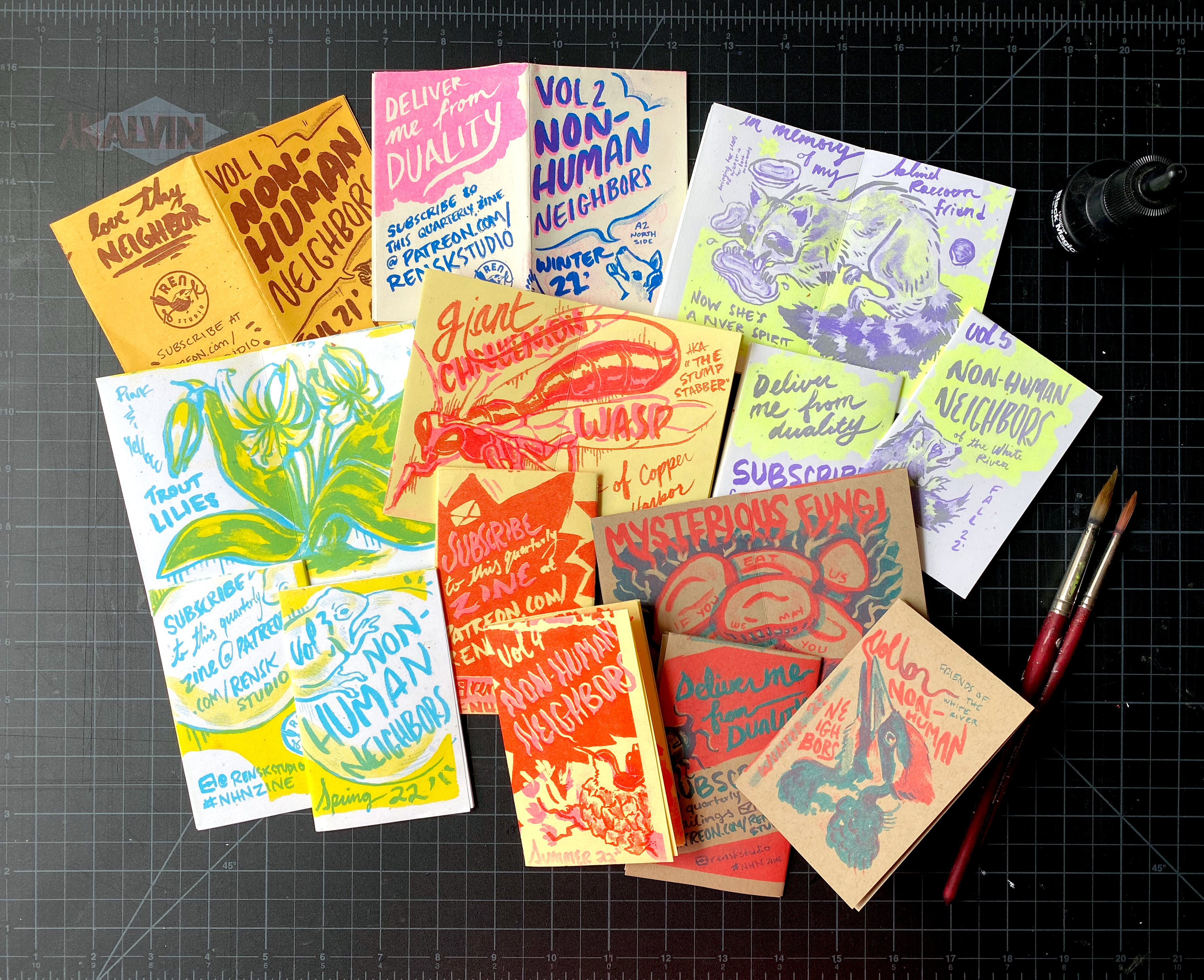 Mini zines on a cutting mat in different colored risograph prints, surrounded by ink brushes and black ink