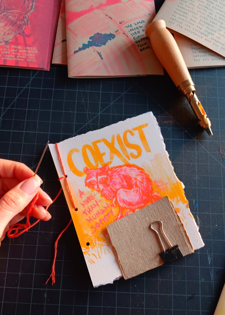 A zine being sewn together on a black background, hand is holding orange thread and needle. THe zine says "Coexist, A more-than-human society" in orange and pink, with a raccoon on the cover. 
