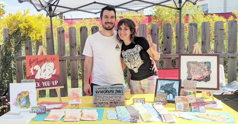 Ren and Mark, artists, vending at a booth outside with zines and prints