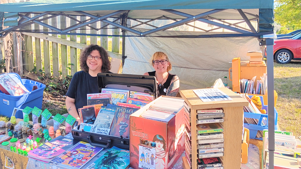 Photo of organizers at their table under a tent. Smiling behind stacks of comics and books.