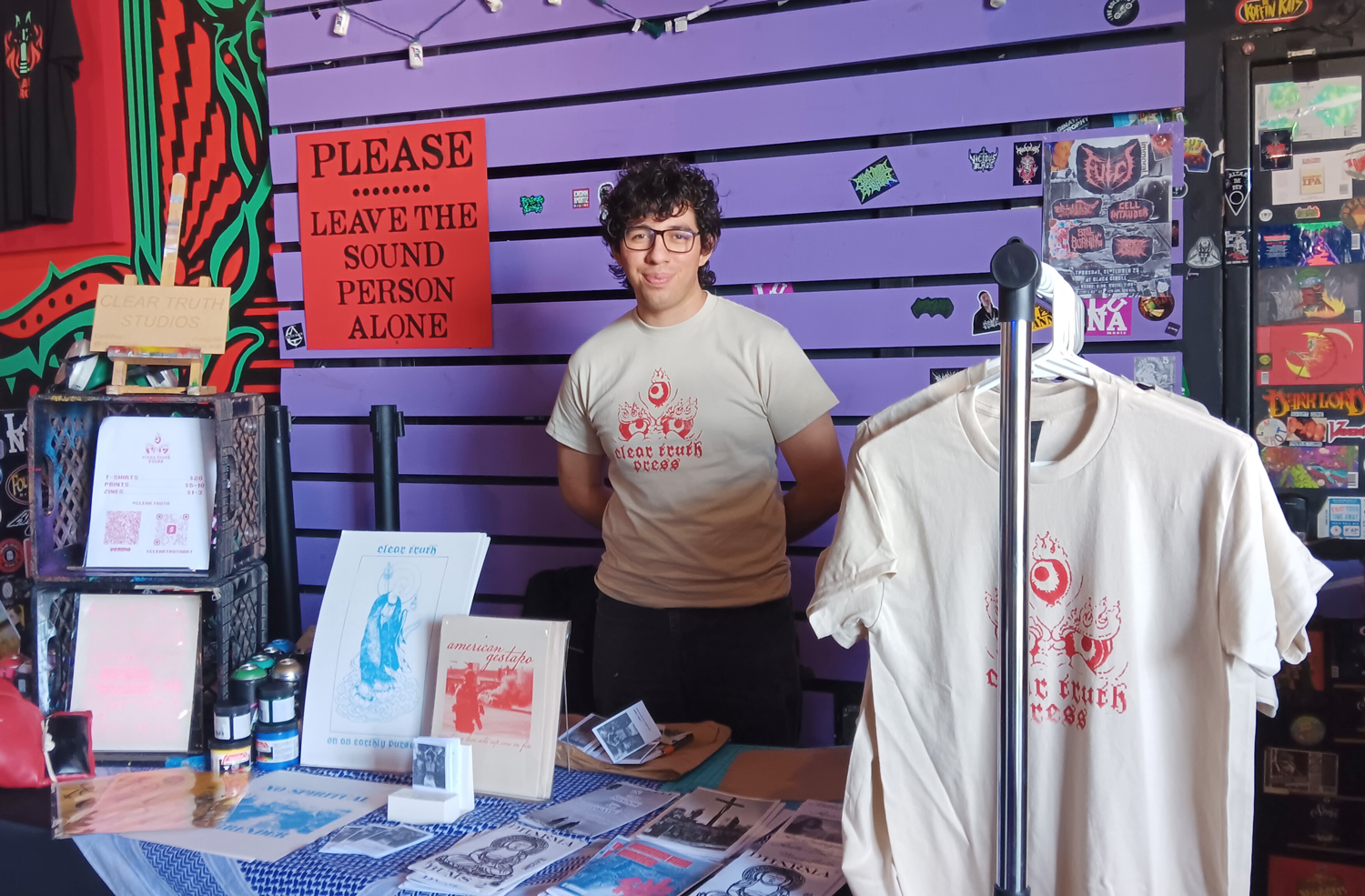 Photo of artist in front of a purple wall. Their table features prints, zines and screen printed shirts