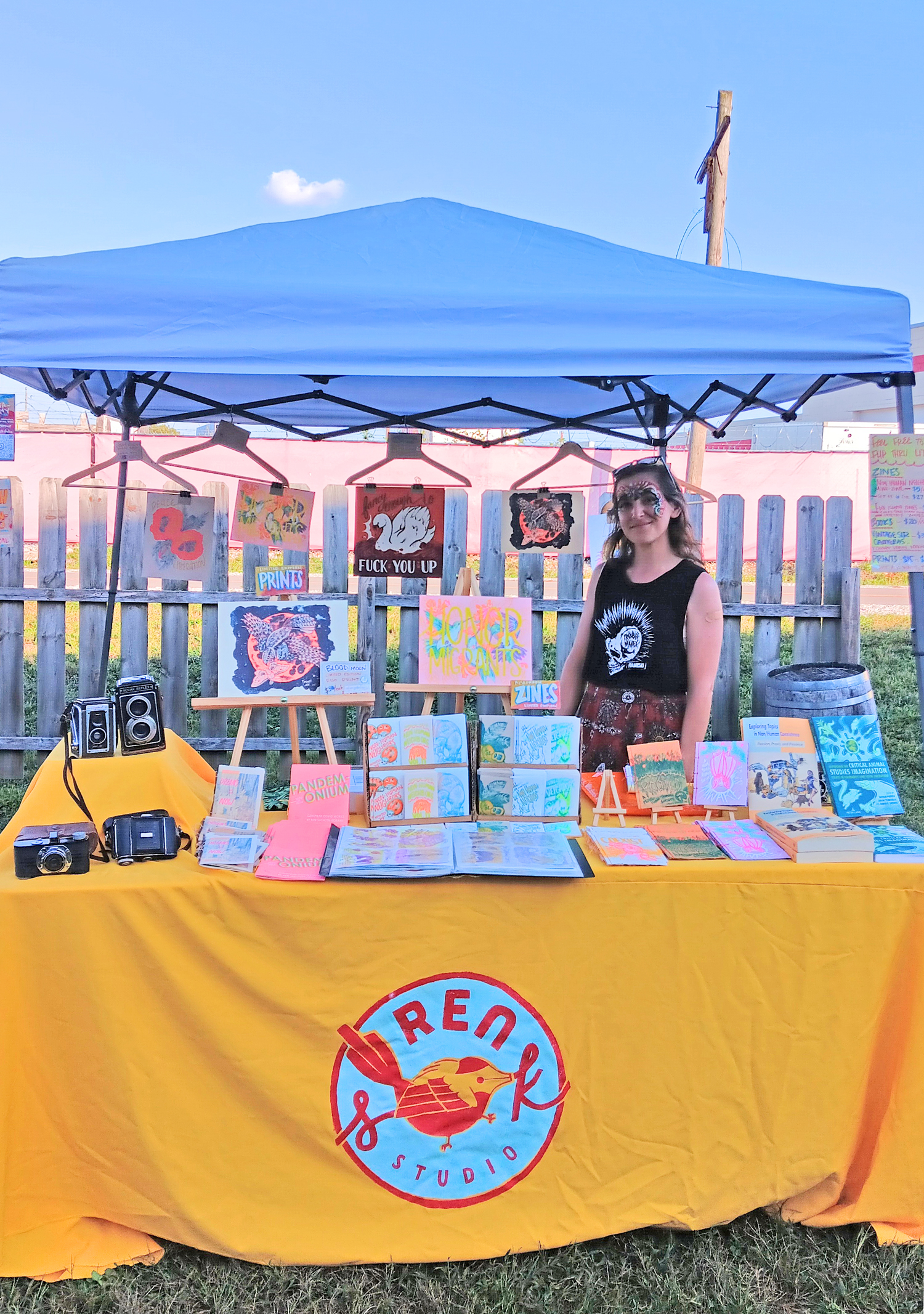 Photo of artist at their booth - table cloth in yellow. ZInes and books and prints and cameras on display