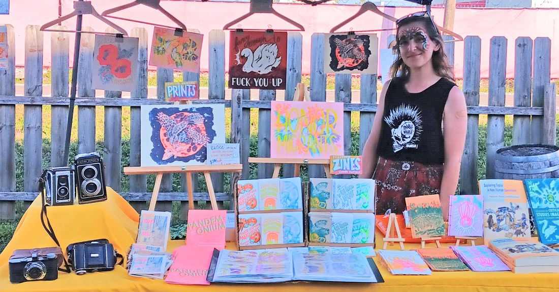 Photo of artist at their booth - table cloth in yellow. ZInes and books and prints and cameras on display. Cropped for wordpress feature size